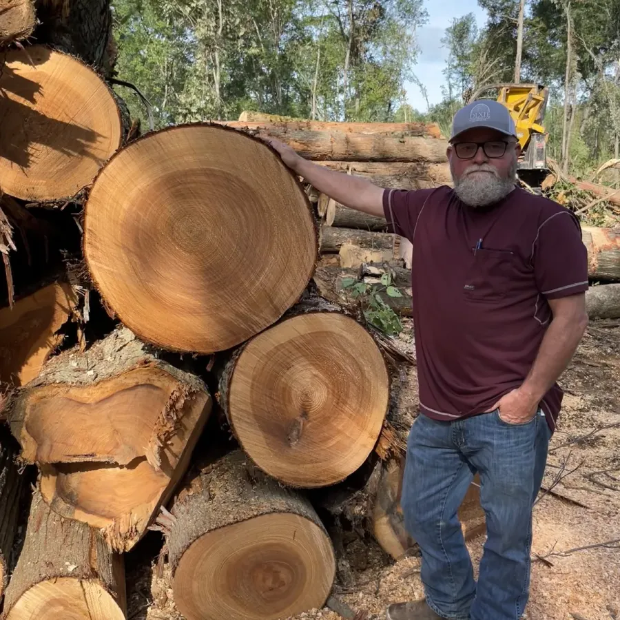 Larry 'Bubba' Hooper standing beside a stack of freshly cut hardwood logs.